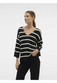 Gestreifter Pullover in Schwarz und Creme mit einem V-Ausschnitt und Dreiviertelärmeln, hergestellt aus einem weichen Stoff, kombiniert mit schwarzen Hosen.