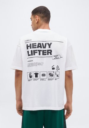 Mand i grønne shorts iført hvid T-shirt med teksten "HEAVY LIFTER" og fitness-tema grafik på ryggen.