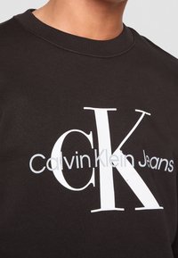 Sweat-shirt noir à col rond avec un grand logo blanc "cK" et "Calvin Klein Jeans" imprimé sur la poitrine.