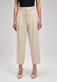 Pantaloni beige a gamba larga con pince, caratterizzati da una texture liscia e una vestibilità sartoriale. Indossati con tacchi beige, abbinati a una maglia nera.