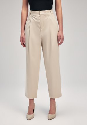 Beige wide-leg trousers with pleats, featuring a smooth texture and tailored fit. Worn with beige heels, complemented by a black top.