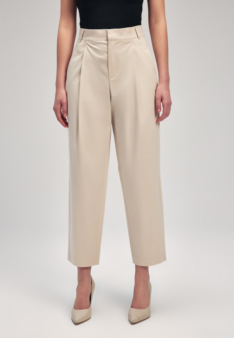 Pantaloni beige a gamba larga con pince, caratterizzati da una texture liscia e una vestibilità sartoriale. Indossati con tacchi beige, abbinati a una maglia nera.