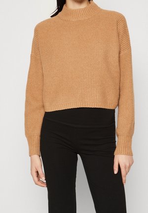 Kobieta ubrana w kameLOWY, teksturowany, krótki sweter dziany, w połączeniu z czarnymi, dopasowanymi spodniami z wysokim stanem na jednolitym tle.