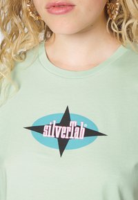 Světle zelené bavlněné tričko s modrým oválným grafickým prvkem, černou hvězdou a textem "silverTab" v růžové barvě. Kulatý výstřih a měkká textura.