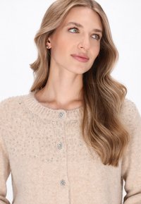 Beiger Cardigan aus weichem Material mit einem strukturierten oberen Bereich, verziert mit klaren Perlenakzenten und glitzernden Knöpfen.