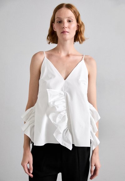 Haut sans manches blanc avec un décolleté en V, des accents en fronces sur les côtés et une texture lisse, associé à un pantalon noir.