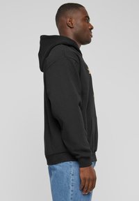 Black hooded sweatshirt with a front pocket, ribbed cuffs, and hem, worn over light blue jeans. Fabric appears smooth and heavy-weight.