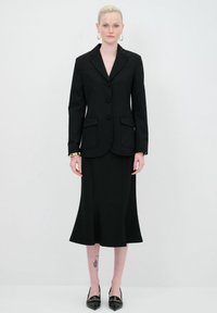 Blazer nero sartoriale con tre bottoni, abbinato a una gonna fitted coordinata. Include tasche laterali e scarpe nere lucide con punta appuntita.