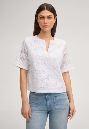 Frau trägt eine weiße Bluse mit kurzen Ärmeln, gemusterten Ausschnitten und dem Schriftzug "JOOP!", kombiniert mit hellblauen Jeans.