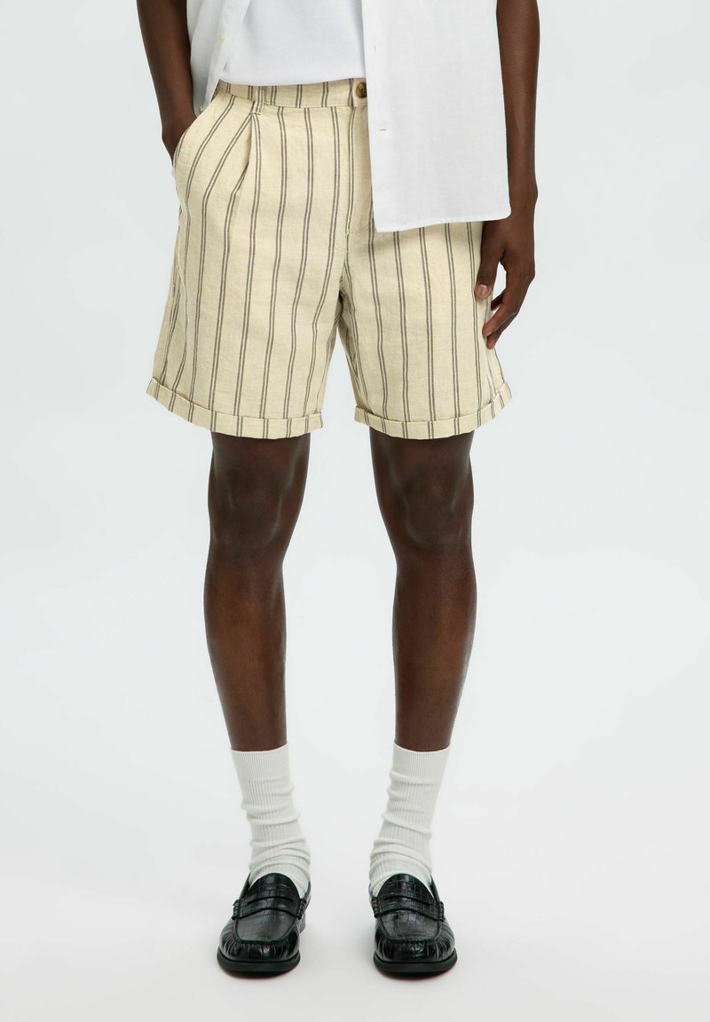 Homme portant un short beige rayé, des chaussettes blanches, des mocassins noirs et une chemise blanche les mains dans les poches sur un fond uni.