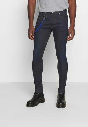 Man wearing dark skinny jeans with blue keychain attached and black leather boots standing against a plain background.