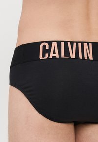 Culottes en coton noir avec une taille élastique douce portant l'inscription "CALVIN" en lettres rose clair. Texture lisse, design ajusté.