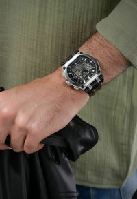 Svart och silver analog klocka med en fyrkantig boett, texturerad svart urtavla och multifunktionella underdials. Hand som håller ett plagg i svart läder.