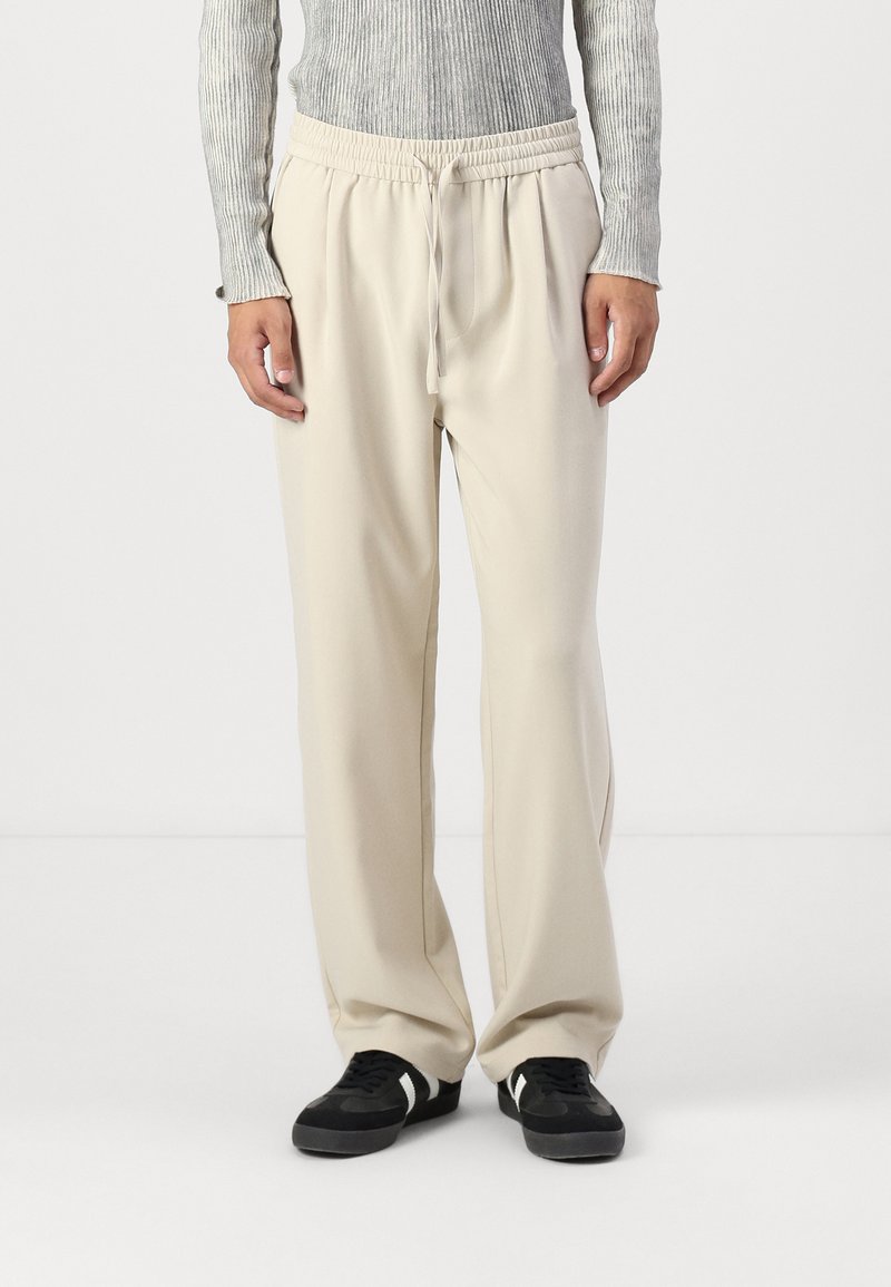 Beige Hosen mit elastischem Bund, lockerer Passform und seitlichen Taschen. Hergestellt aus weichem Stoff, mit einer Kordelzug-Details und weitem Beindesign.