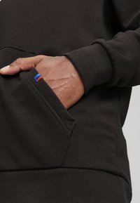 Dark green sweatshirt with a ribbed cuff, featuring a kangaroo pocket and a small red and blue logo accent on the pocket edge.