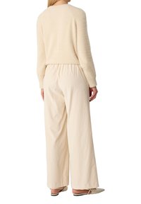 Frau trägt einen cremefarbenen, flauschigen Pullover, lockere hoch-taillierte, weite Hosen und helle, spitze Slingback-Schuhe, steht mit dem Rücken zur Kamera.