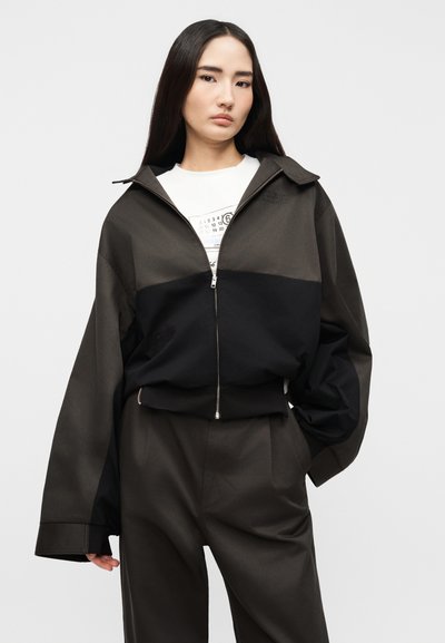 Γυναίκα με μακριά σκούρα μαλλιά που φοράει φαρδιά διχρωμία σε μαύρο και σκούρο γκρι μπουφάν με φερμουάρ και ταιριαστό σκούρο γκρι παντελόνι, σε λευκό φόντο.