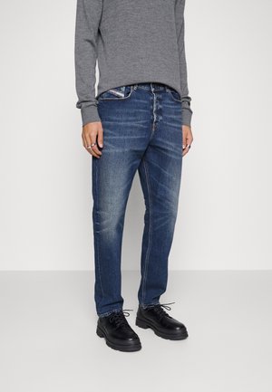 Personne portant un jean bleu délavé, une chemise grise à manches longues, des chaussures noires à lacets et des bagues en argent aux deux mains.
