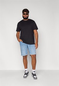 Schwarzes T-Shirt, hellblaue Jeansshorts mit umgeschlagenen Säumen und schwarz-weiße Sneakers. Die Figur trägt dunkle Sonnenbrillen und steht lässig.
