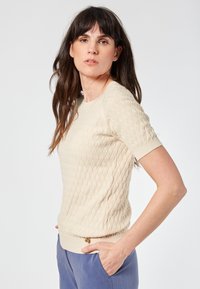 Beige knitted sweater with a textured diamond pattern, short sleeves, and a ribbed hem. Accompanied by blue trousers.