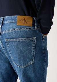 Dżinsy w średnim niebieskim kolorze z delikatnym przetarciem. Posiadają skórzaną naszywkę z napisem "Calvin Klein Jeans", nitami i tylnymi kieszeniami.