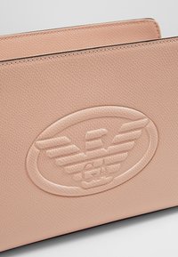 Růžová texturovaná kožená taška s vyraženým logem s křídly a kruhovým rámem. Hladký povrch s precizním prošív áním podél okrajů.