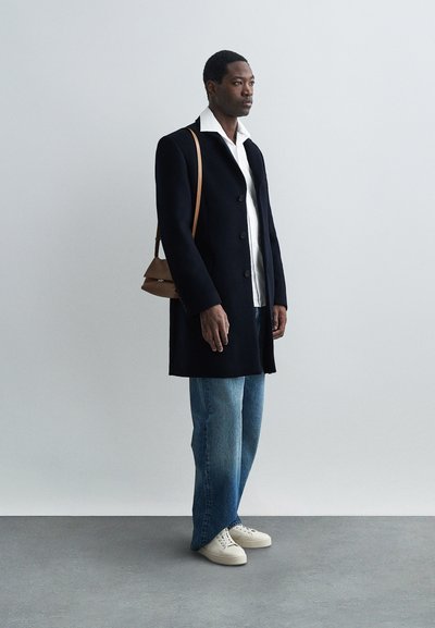 Navy coat with button closure, white shirt underneath, light blue wide-leg jeans, and white sneakers. Brown crossbody bag on shoulder.