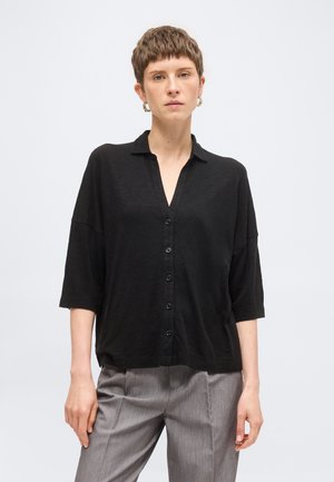 Mujer con el pelo corto que lleva una camisa negra suelta abotonada y pantalones grises, de pie frente a un fondo liso y claro.