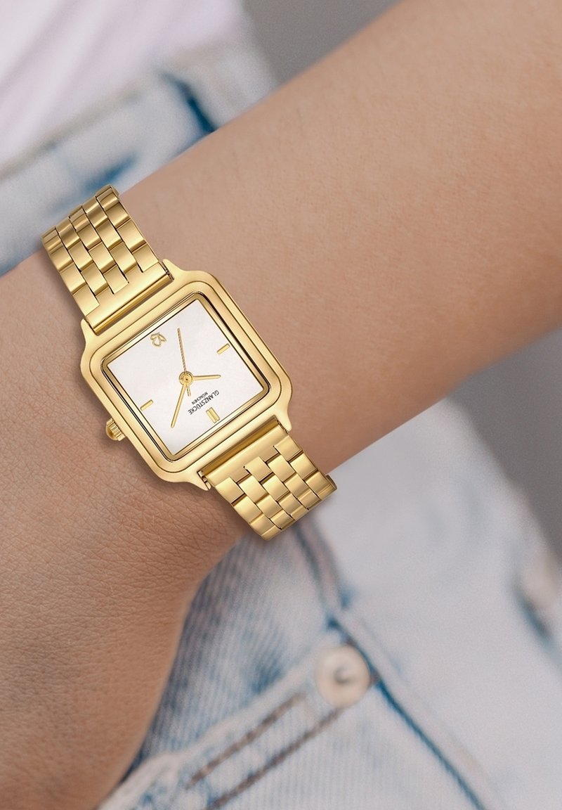 Kuldtooni ruudukujuline kell valge näo, kuldsete käte ja lingikettide disainiga, puhkab heledal naha tooni randmel.