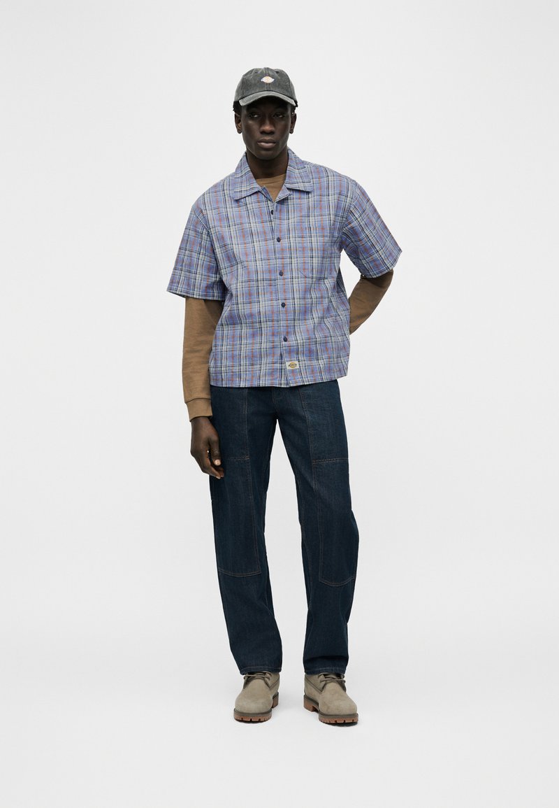 Uomo con cappellino grigio, camicia a maniche corte a quadri blu e viola sopra una camicia marrone a maniche lunghe, jeans scuri e stivali beige, in piedi contro uno sfondo bianco.