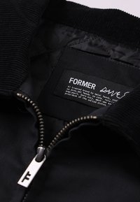 Μαύρο μπουφάν με γιακά από κορδόνι, με μεταλλικό φερμουάρ και εσωτερική ετικέτα που γράφει "FORMER" με επιπλέον κείμενο. Λείο ύφασμα.