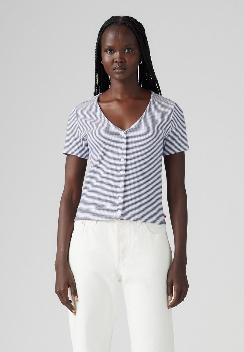 Chemise à rayures bleues et blanches à manches courtes avec un col en V et un bouton, fabriquée dans un tissu léger, associée à un pantalon blanc.
