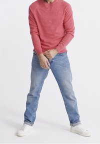 Homme portant une chemise corail à manches longues, un jean bleu clair et des baskets blanches, debout jambes écartées, ajustant son jean avec les mains.