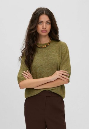 Une femme porte un pull à manches courtes vert olive, un pantalon marron et un collier de perles devant un fond uni et clair.