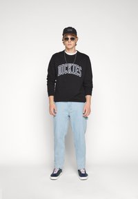 Black sweatshirt with "DICKIES" print, light blue jeans, and navy sneakers. Accessorized with sunglasses and a black cap.
