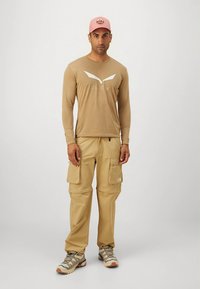 Beige langærmet skjorte med hvide detaljer og logo, kombineret med cargobukser i khaki. Grå vandresko med lilla og cremefarvede detaljer. Lyserød kasket.