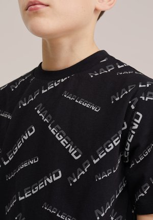 Młoda osoba ubrana w czarny T-shirt z powtarzającym się szarym napisem "NAP LEGEND" na jednolitym tle.