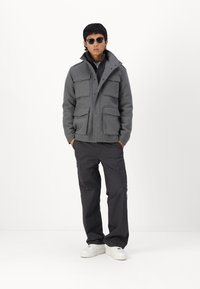 Chaqueta gris texturizada con cuatro bolsillos en la parte frontal, cierre de cremallera y cuello alzado. Combinada con pantalones cargo oscuros y zapatillas blancas. Se usan gafas de sol negras.