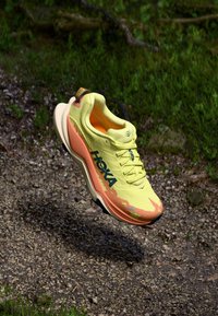 Chaussure de sport jaune avec une surface texturée, des accents dégradés orange, un logo proéminent et une semelle en caoutchouc, flottant au-dessus d'un chemin de gravier.