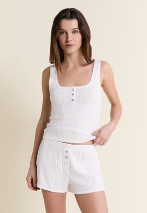 Junge Frau trägt weißes, geripptes Tanktop und passende Shorts mit Knopfdetails, steht vor einem schlichten beigen Hintergrund.