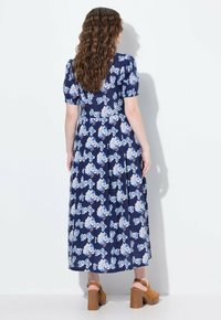 Květinové maxi šaty v námořnické modré barvě s modrým a růžovým květinovým vzorem. Mají krátké nadýchané rukávy a splývavou sukni. Jsou nošeny s hnědými sandály na silném podpatku.