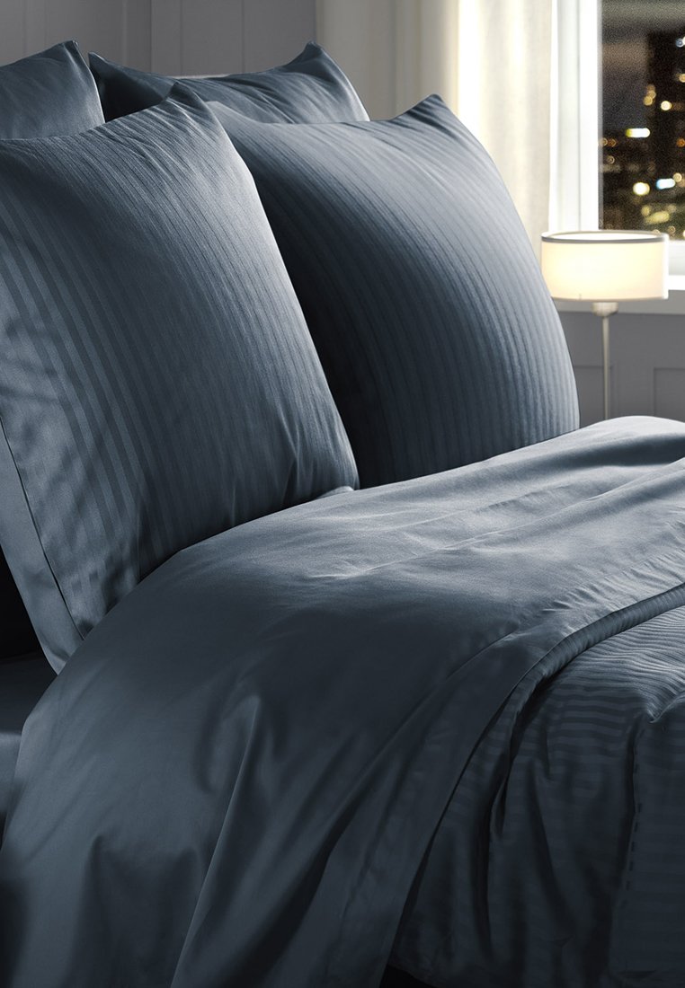 Dwie duże niebieskie poduszki w paski oraz pasujący niebieski narzuta na łóżku, a w tle świecąca lampa i okno.