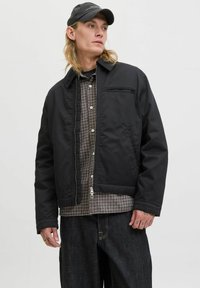 Jeune personne avec de longs cheveux blonds portant une casquette noire, une veste zippée noire, une chemise à carreaux et un jean foncé sur un fond blanc.