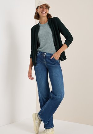 Groene fleecejack met rits over een lichtgrijs T-shirt, gecombineerd met blauwe hoge taille jeans en crèmekleurige sneakers, tegen een neutrale achtergrond.