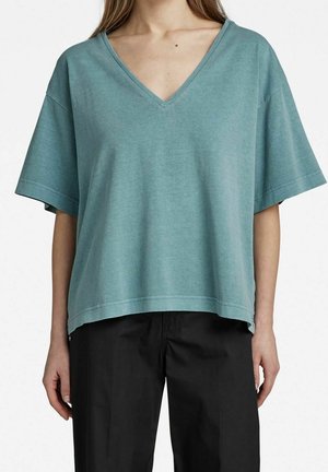 Turkusowy oversize'owy T-shirt z dekoltem w serek, wykonany z miękkiej tkaniny, posiada szerokie krótkie rękawy i prosty dół. W połączeniu z czarnymi spodniami.