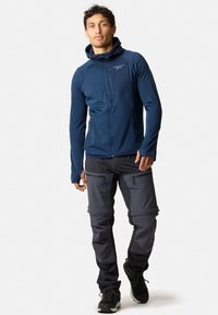 Marineblauwe zip-up jas met een getailleerd ontwerp, textuurstof en capuchon, gecombineerd met grijze cargo-broek met ritsen en zwarte wandelschoenen.