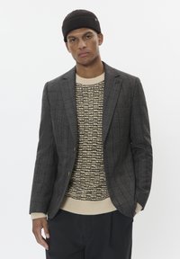 Blazer de mezcla de lana gris con un sutil patrón de cuadros, diseño de un solo botón, solapas muesca y botones en tono dorado, sobre un suéter beige de punto.