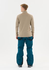 Beige longsleeve top met hoge kraag, gecombineerd met blauwe skibroek met zwarte accenten en versterkte zomen. Model van achteren gezien.