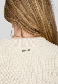 Pull en tricot crème avec une texture côtelée. Doté d'un large col et d'une petite étiquette en métal argenté à l'arrière du col.