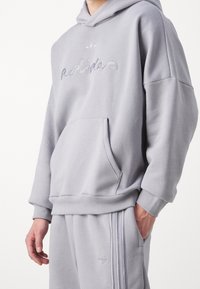 Person som bär en ljusgrå Adidas hoodie och matchande träningsbyxor med logotyp och signatur broderad på bröstet och ränder på byxorna.